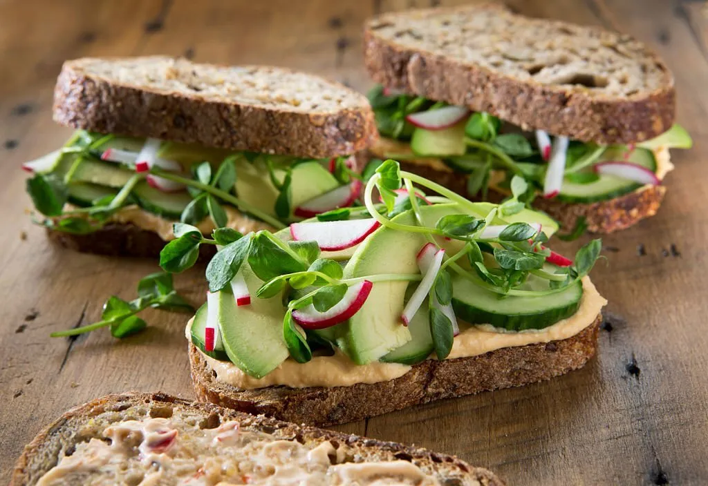 Three healthy vegetarian sandwich's on a rustic wood background. Sandwich's are on multigrain bread with roasted red pepper hummus , radish's ,cucumber , pea shoots and avocado.